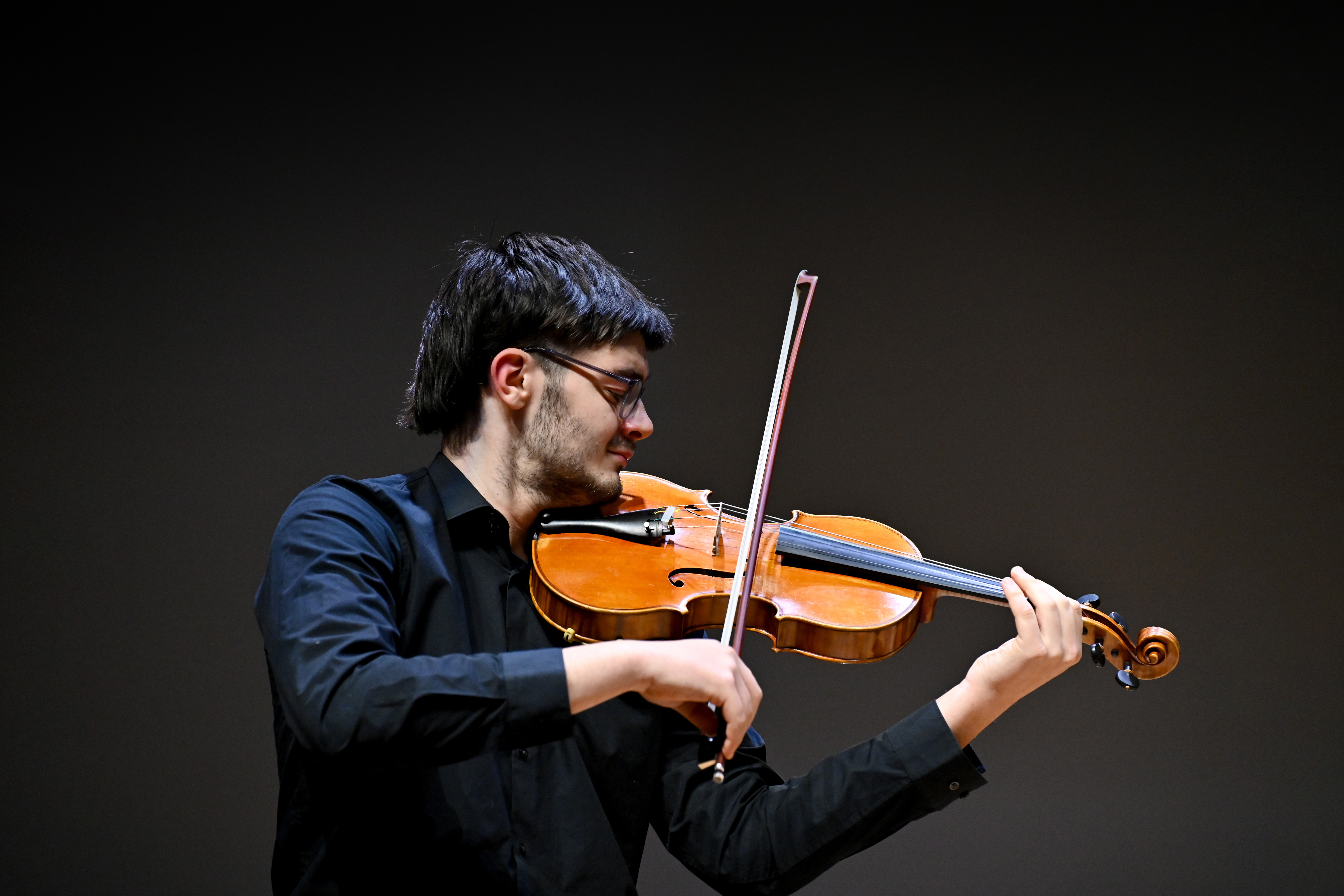 A male student wearing a black shirt, playing the violin in a performance.
